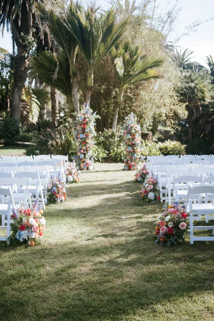Outdoor wedding aisle lined with white chairs and colorful floral arrangements beneath tall palm trees