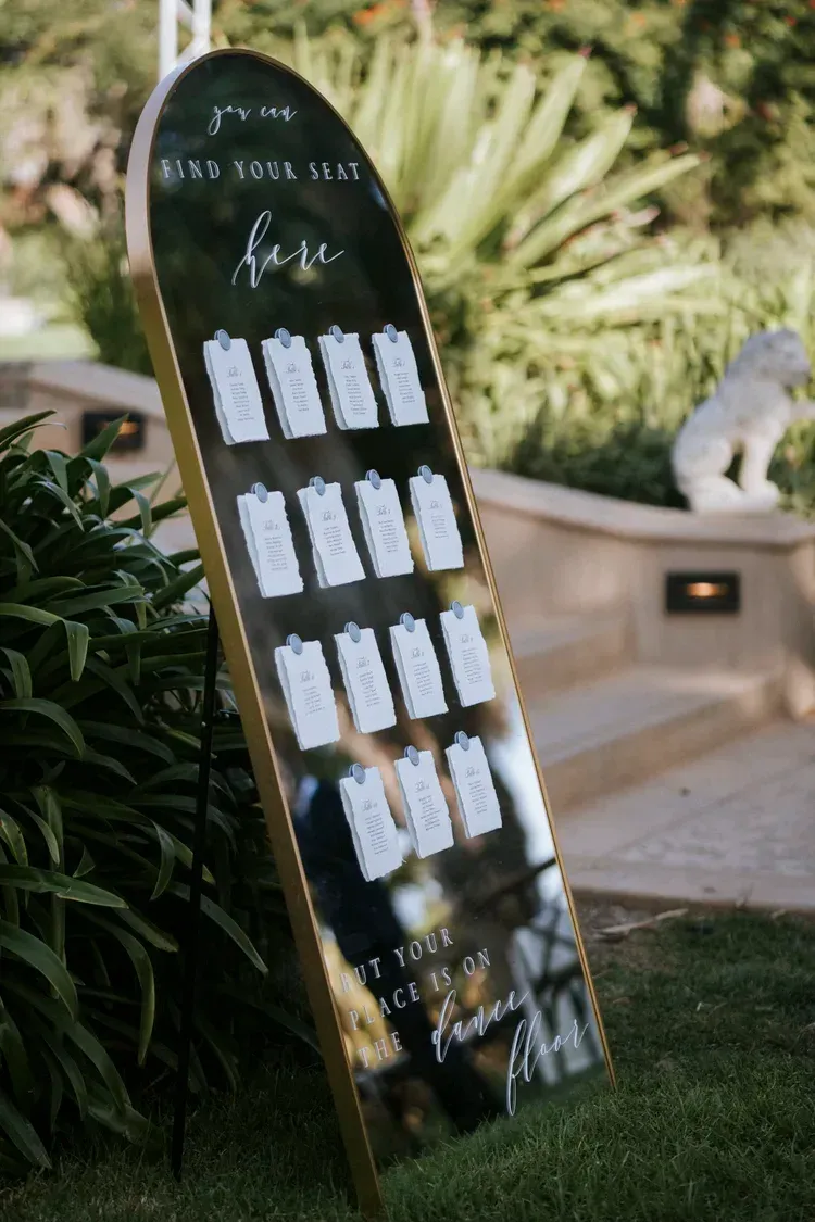 Black outdoor seating chart sign with white cards, leaning beside greenery on a lawn.