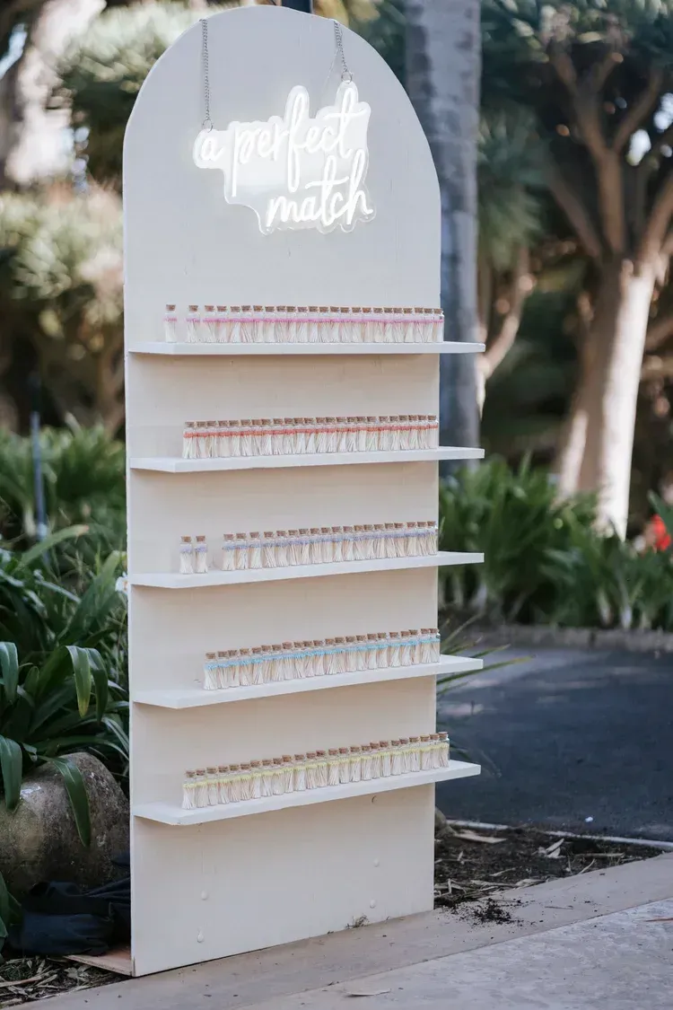 White outdoor display stand with neon sign and rows of small items on shelves, set in a garden path.