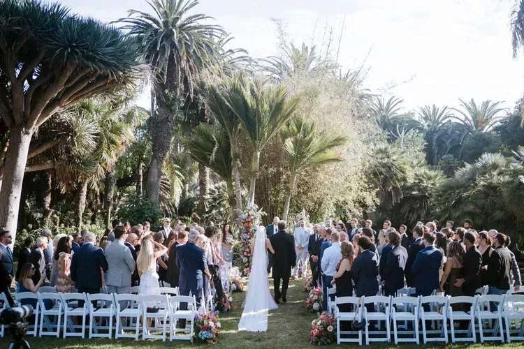 Outdoor wedding ceremony among palm trees, guests standing beside white chairs in a garden setting