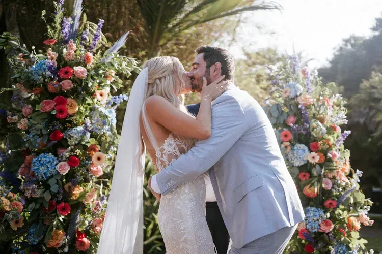 Bride and groom kiss under floral arch in a sunny garden ceremony