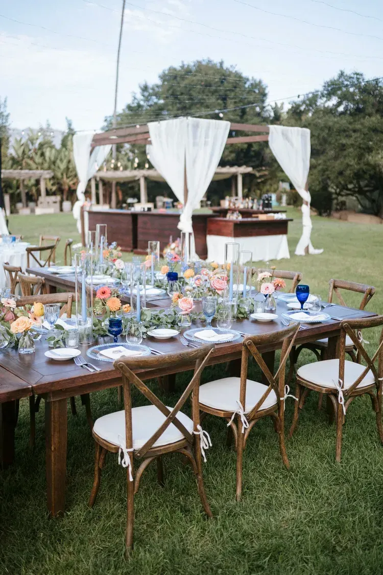 Outdoor banquet table with white chairs and floral centerpiece beside a white-draped canopy bar in a grassy yard