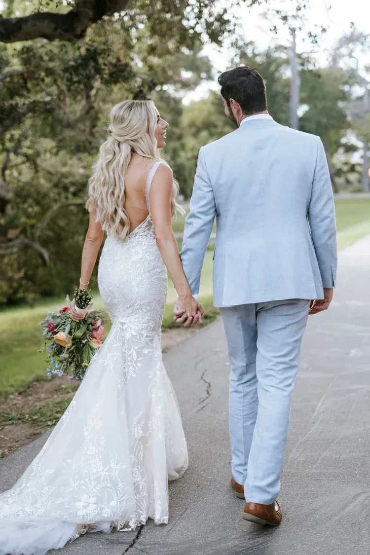 Couple in formal attire holding hands while walking down a tree-lined road outdoors.