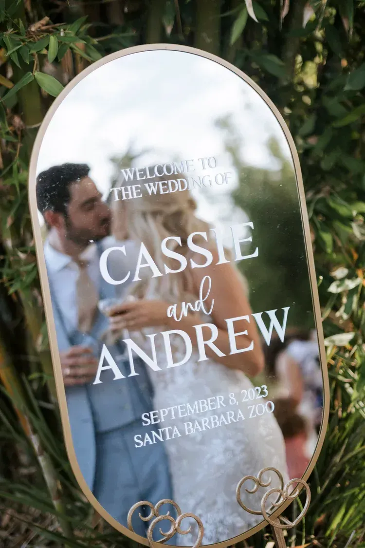 Oval wedding mirror sign reading “Cassie and Andrew” with a couple’s reflection in the background.