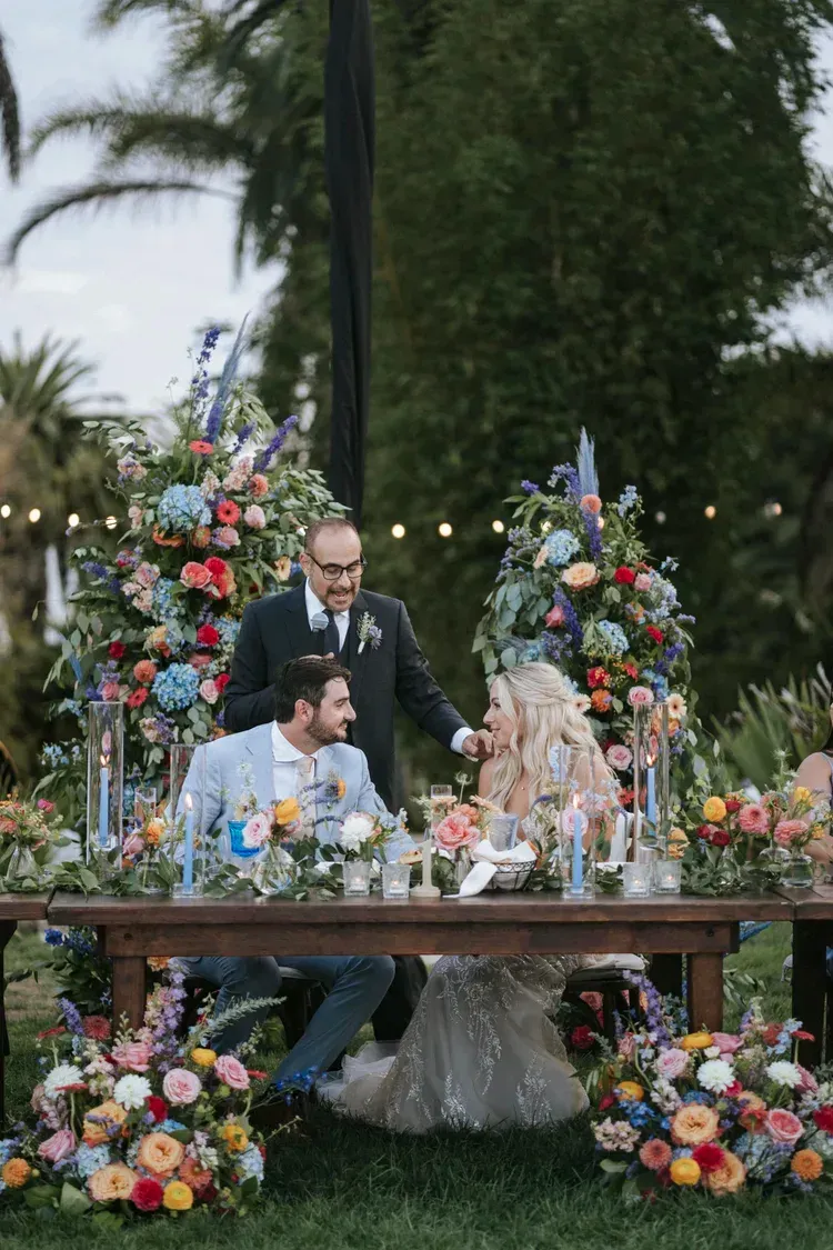 Three people at a flower-filled outdoor wedding table, with a seated couple and a standing man behind them.