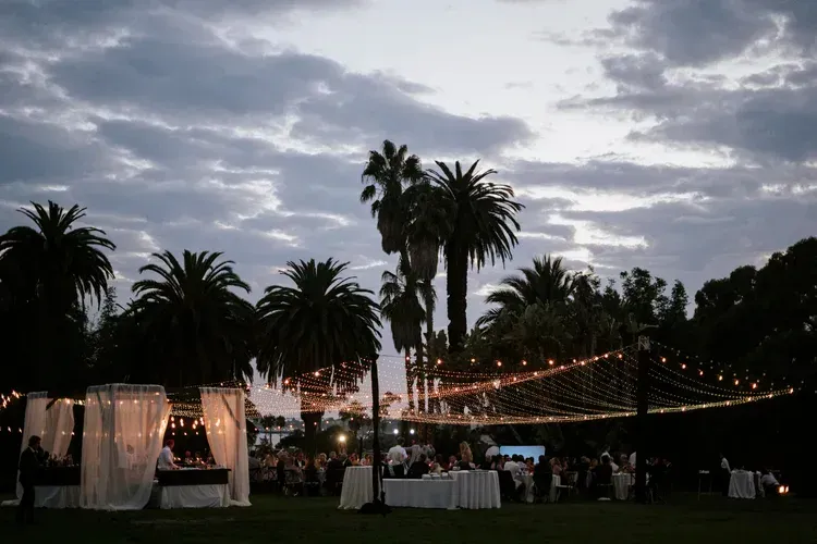 Outdoor evening event with palm trees, string lights, and white tents under a cloudy sky
