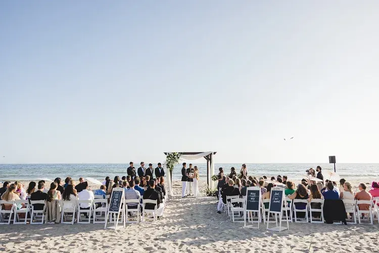 Beach wedding ceremony with guests seated in white chairs under a small arch by the ocean