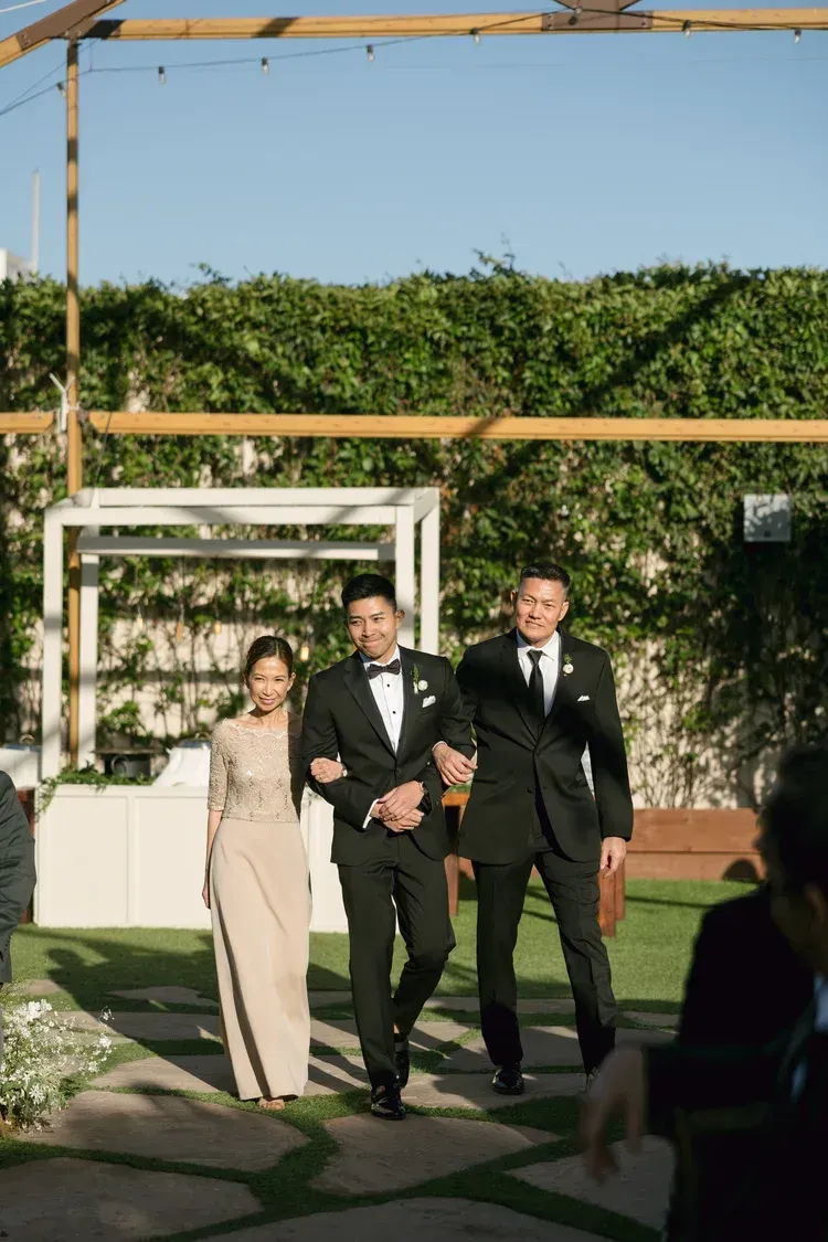 Three people in formal attire walking down an outdoor aisle at a sunny garden ceremony