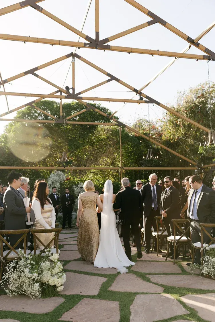 Outdoor wedding ceremony under a wooden pergola with guests gathered around the bride and groom