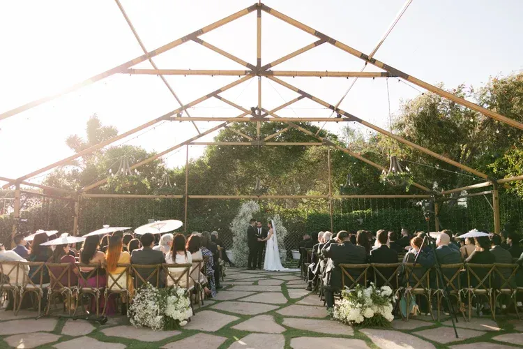 Outdoor wedding ceremony under a wooden canopy, with guests seated on both sides of an aisle leading to the bride.