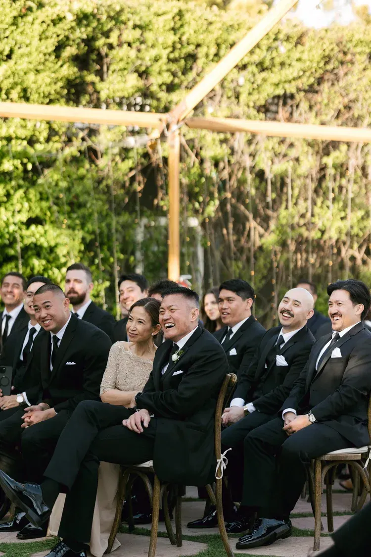 Group of formally dressed guests seated outdoors under a canopy, watching an event.