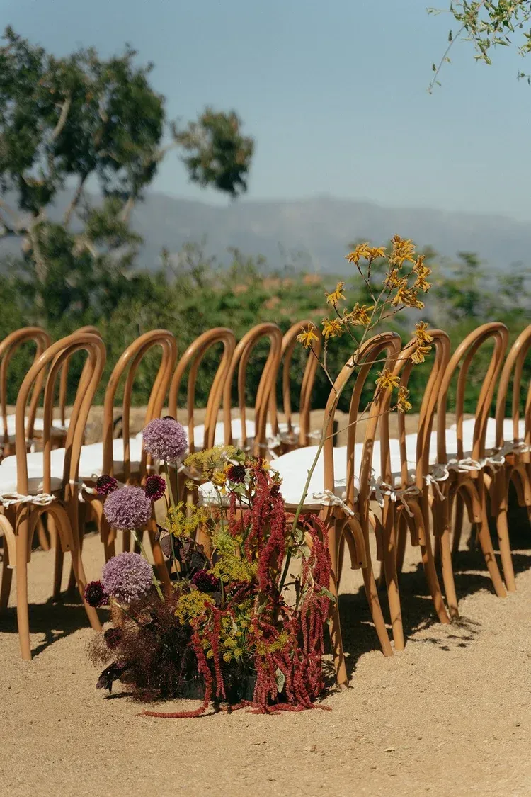 Rows of wicker chairs outdoors with colorful flower arrangements in front of a blurred landscape.