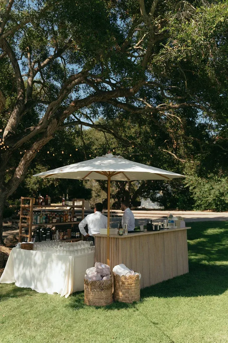 Outdoor bar setup under a large tree with white canopy and beige tablecloths on a grassy lawn.