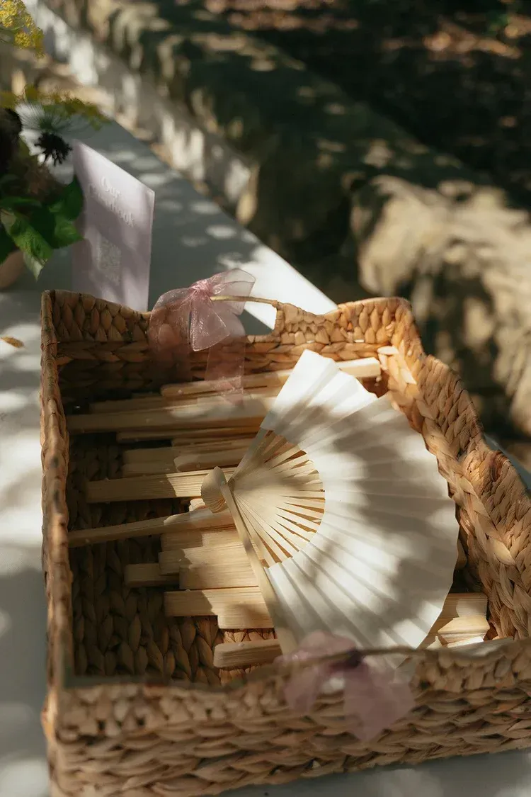 Wicker basket with folded paper fan and small cloth items in dappled sunlight outdoors