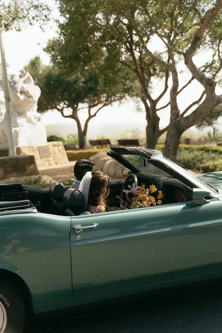 Couple in a green convertible decorated with flowers, driving along a tree-lined road in daylight