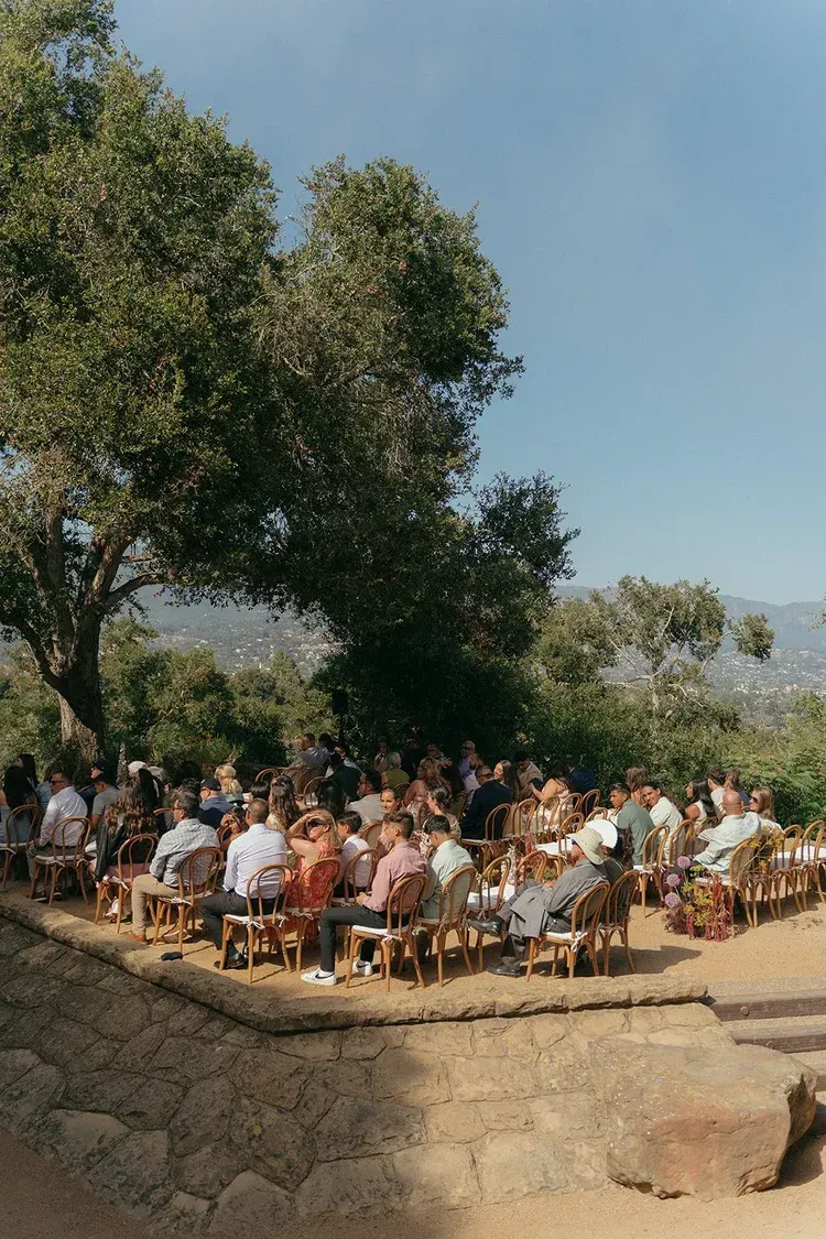 Outdoor gathering of people seated in a circle under trees on a sunny hillside