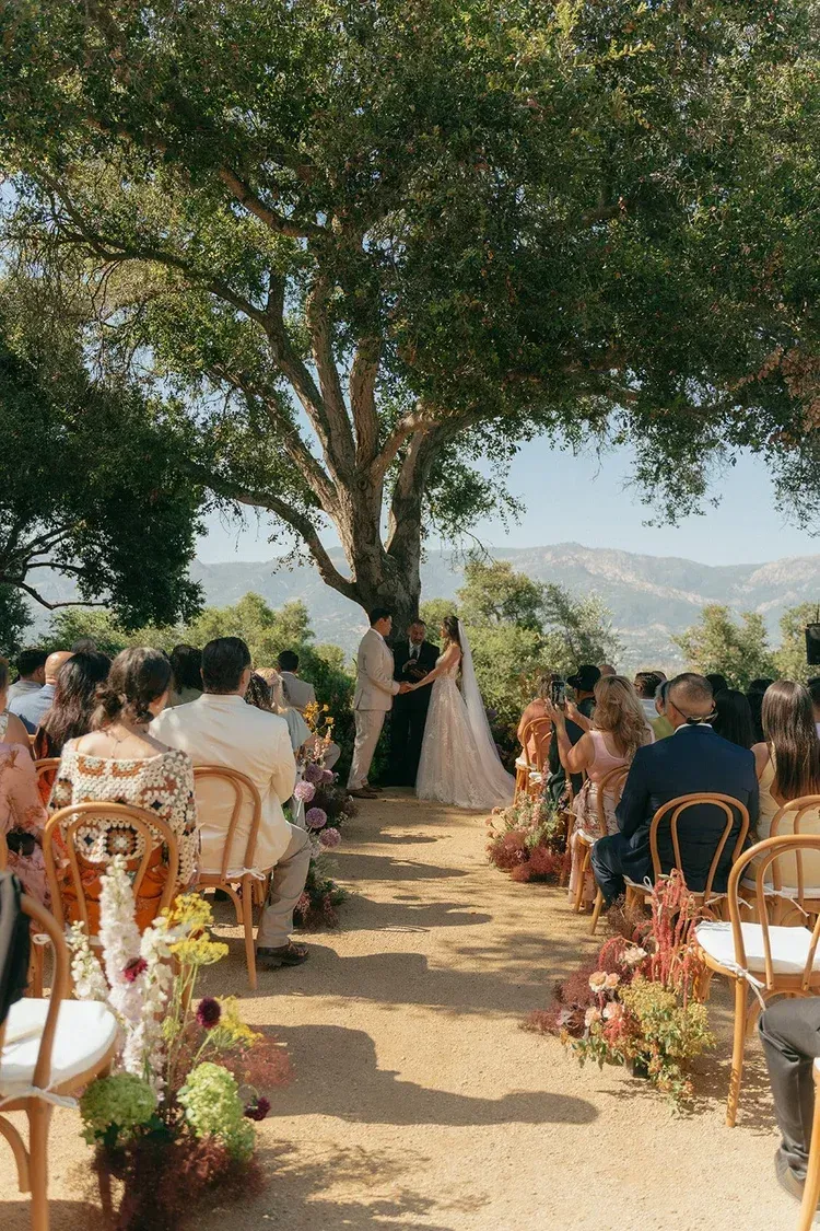 Outdoor wedding ceremony beneath a large tree, with guests seated along a flower-lined aisle.