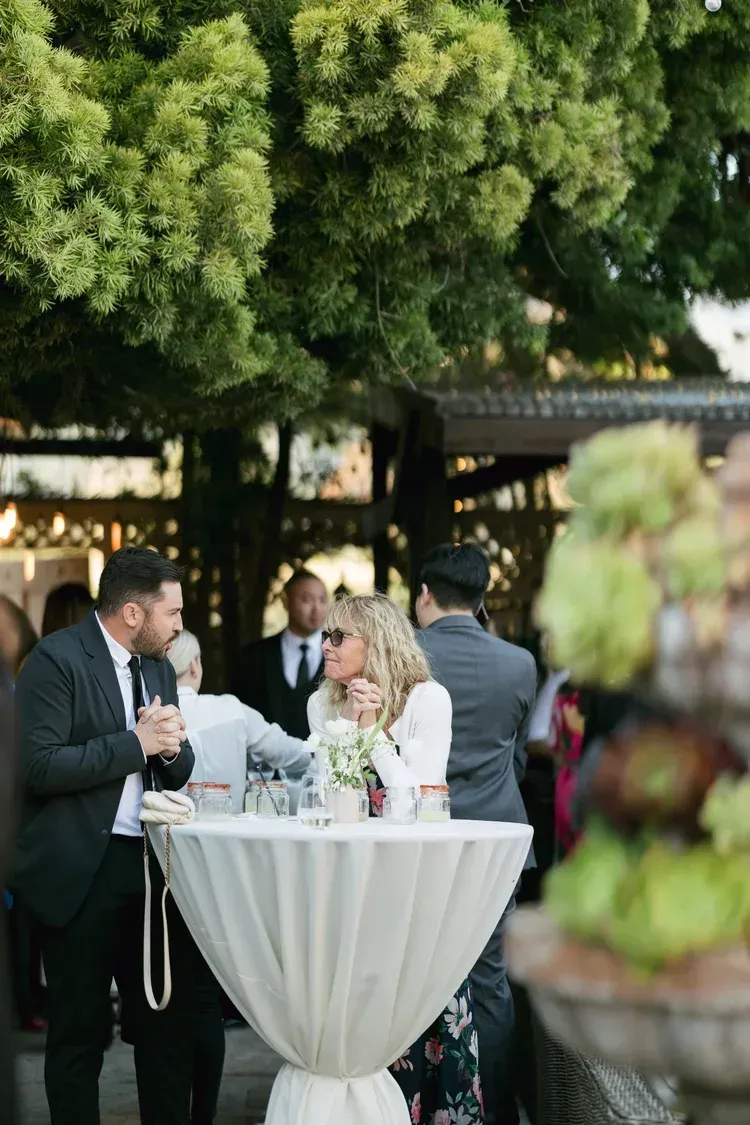 Guests chatting at a cocktail table during an outdoor wedding reception under lush greenery