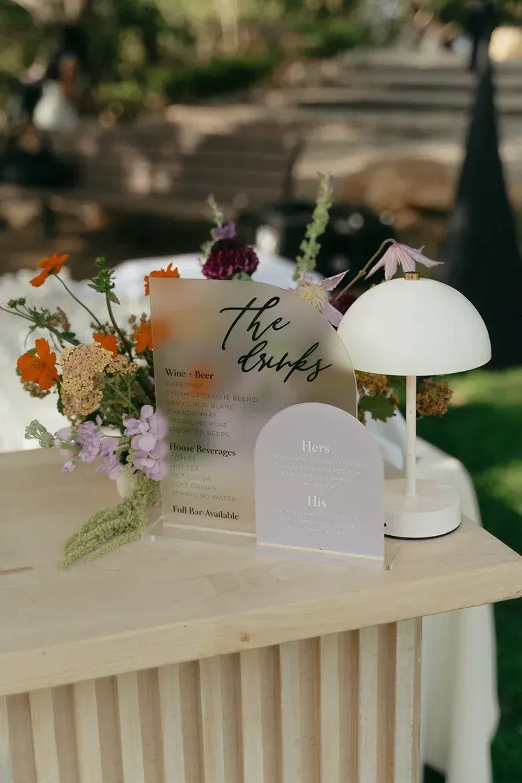 Elegant floral table centerpiece with handwritten sign and white lamp on a wooden table outdoors