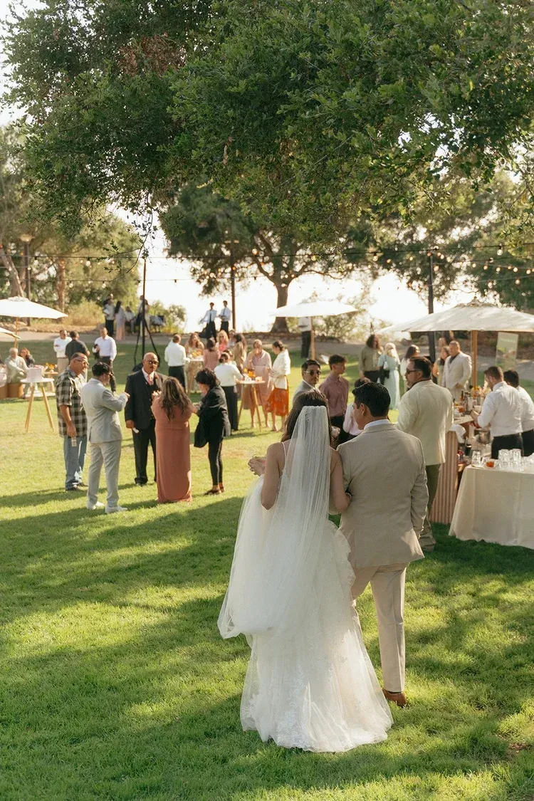 Bride and groom walking through a sunny garden wedding reception with guests nearby