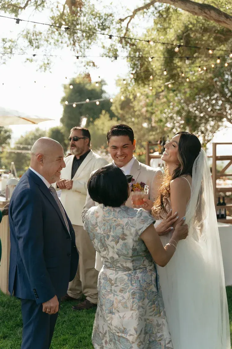 Wedding group exchanging a toast outdoors under trees, with the bride in a white veil and guests gathered nearby