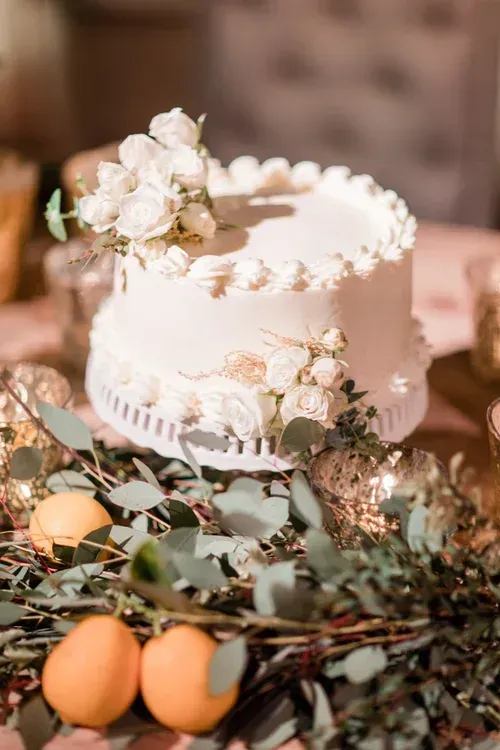 White tiered cake with flowers, surrounded by greenery and oranges on a table