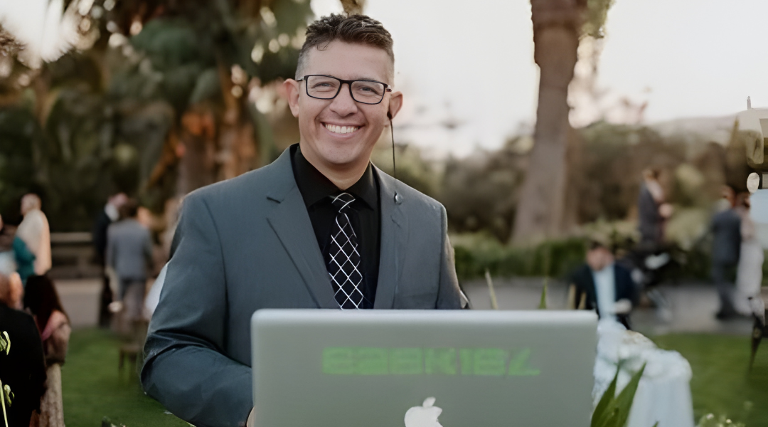 Smiling man in a blue suit standing outdoors behind a laptop at an event