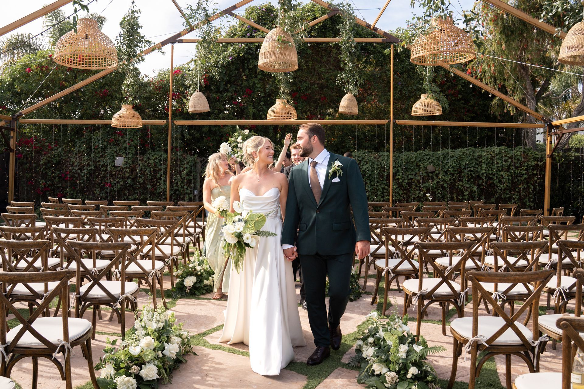 Bride and groom walking down an outdoor wedding aisle beneath hanging floral baskets