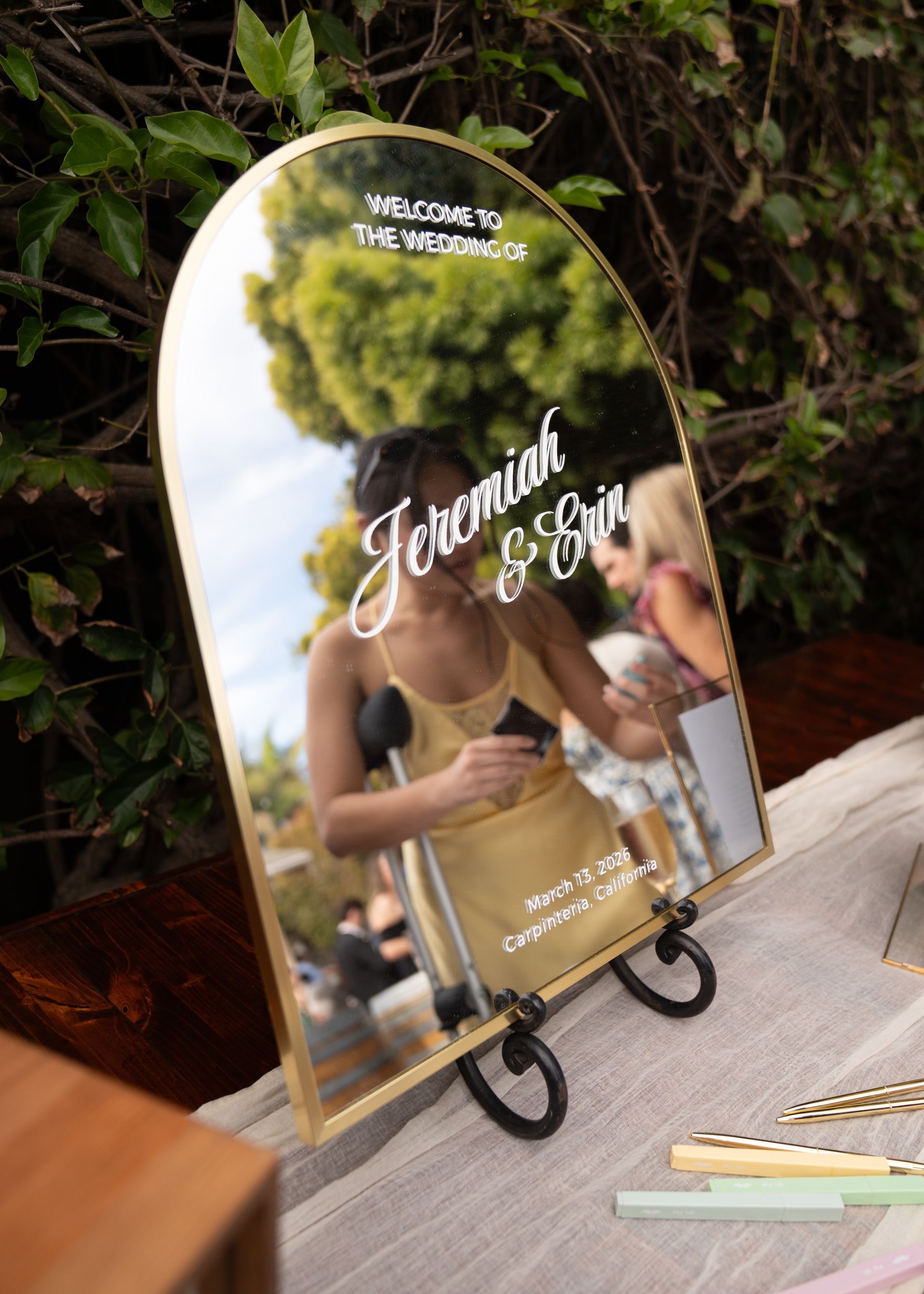 Mirror sign with white script on a stand, reflecting greenery and a person holding a champagne glass.