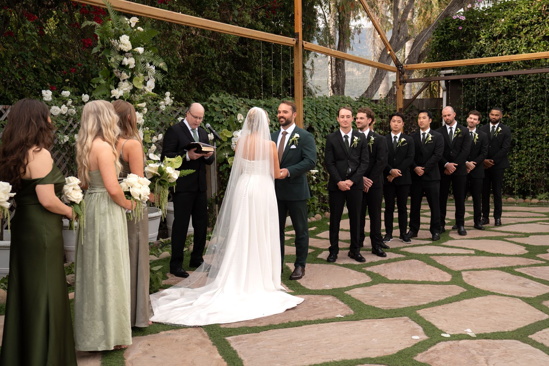 Wedding ceremony in a garden, bride in white facing groom and groomsmen in black suits