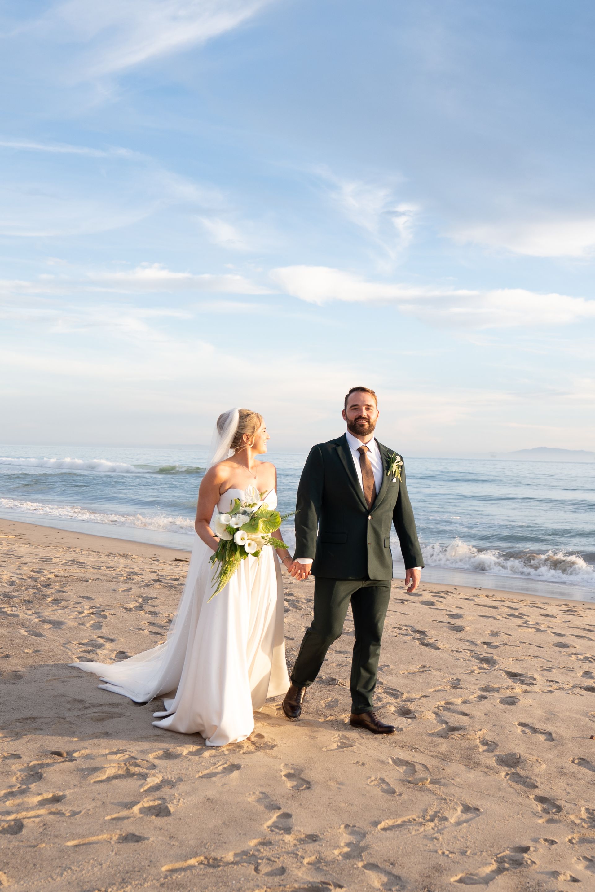 Bride and groom walking hand-in-hand on a beach at sunset, bride in white dress holding bouquet