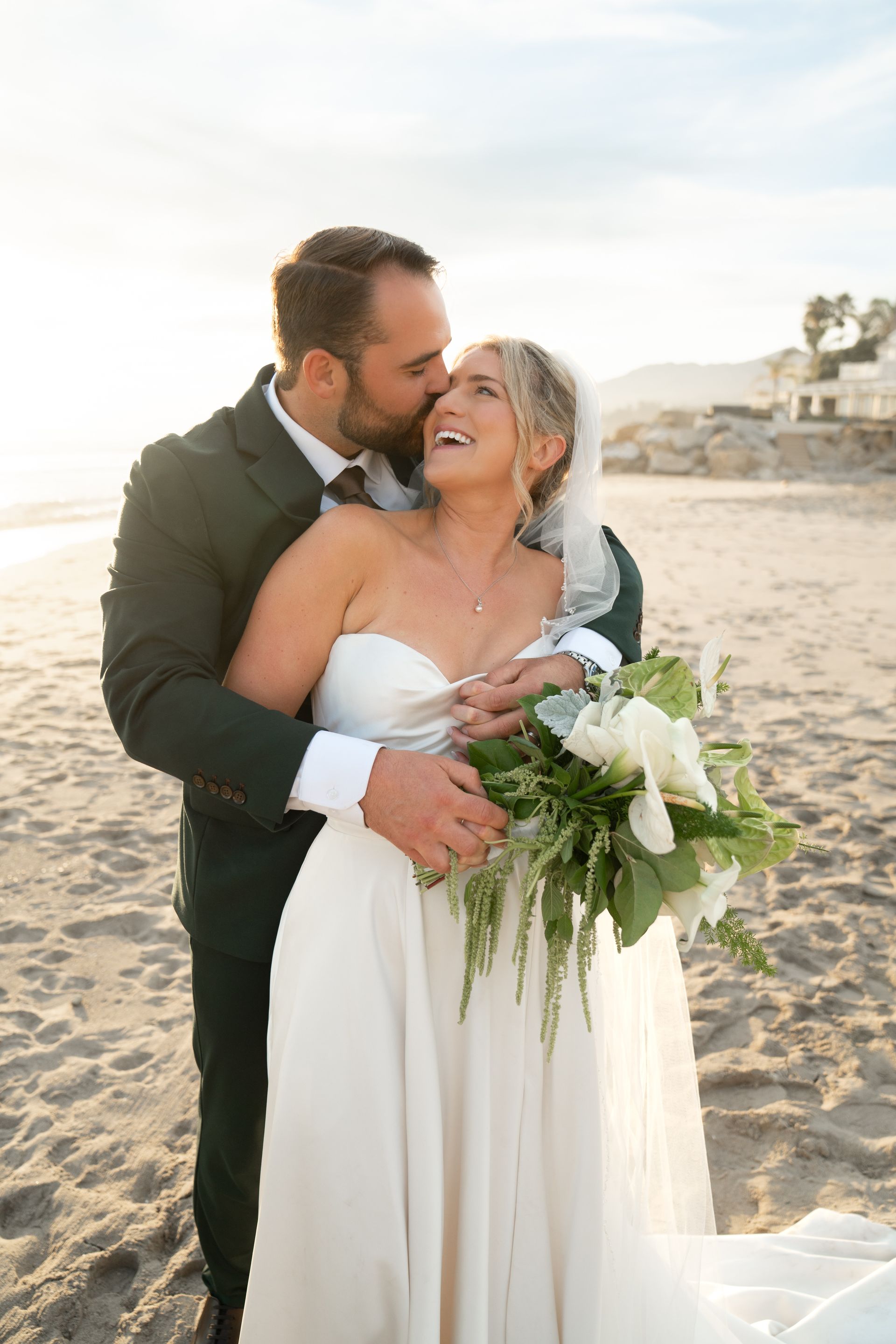 Bride and groom kissing on a sunny beach, holding a white bouquet.