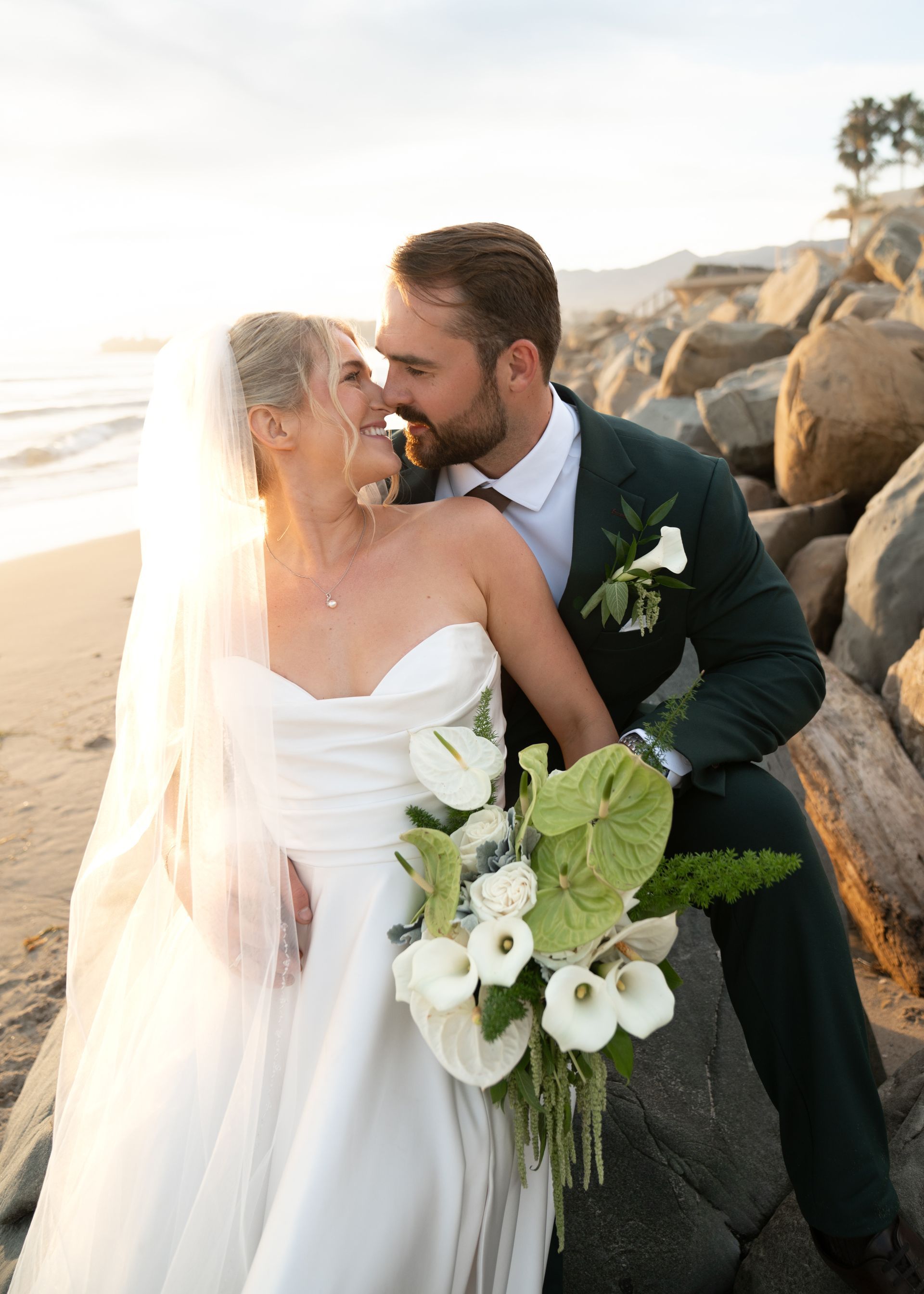 Bride and groom embracing on a rocky beach at sunset, bride holding a white flower bouquet.