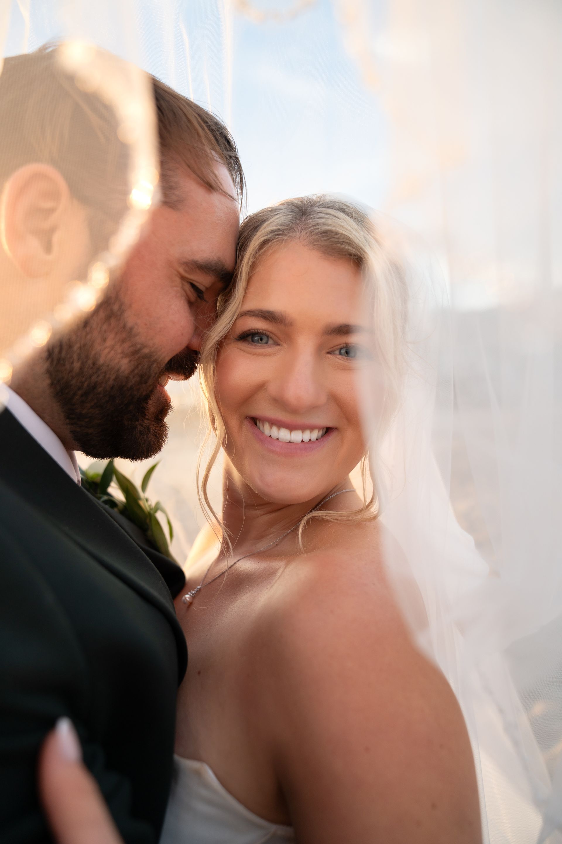 Smiling bride and groom in a warm, backlit close-up embrace outdoors