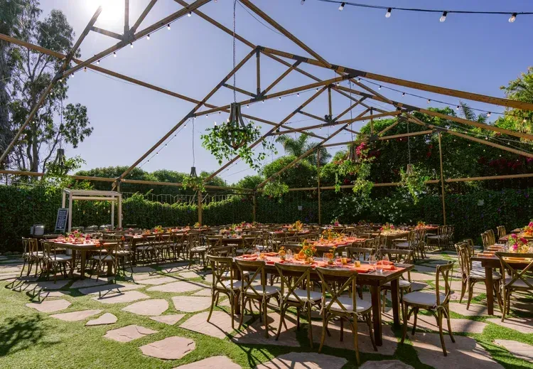 Outdoor dining area with wooden tables under a metal frame canopy and string lights, surrounded by greenery