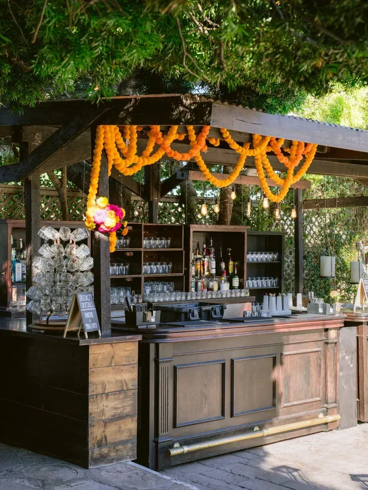 Outdoor bar decorated with orange marigold garlands, under a leafy canopy, with shelves of bottles and glassware