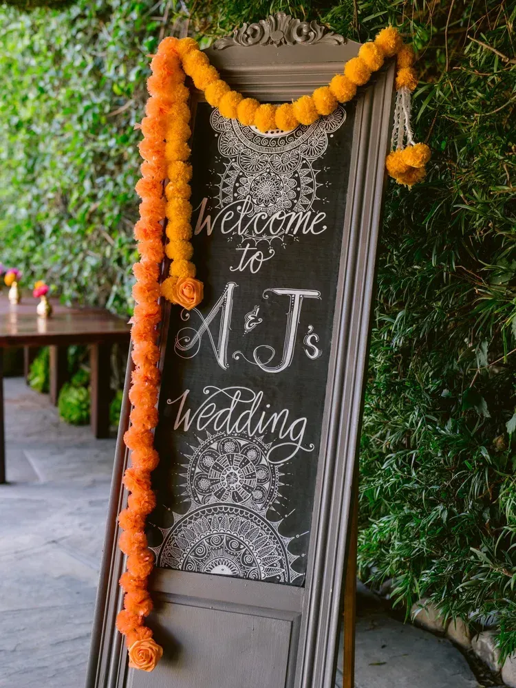 Chalkboard wedding sign with orange marigold garlands reading “Welcome to N.J.’s Wedding”
