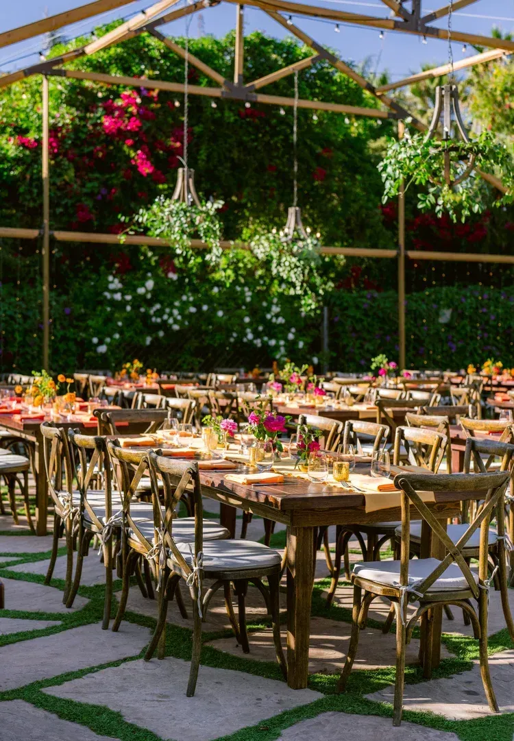 Outdoor dining setup with bamboo tables and chairs under a shaded trellis, surrounded by lush greenery and flowers
