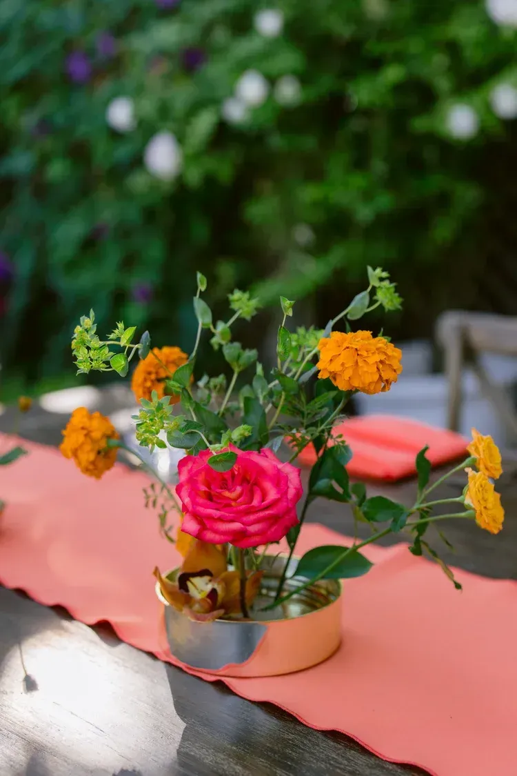 Pink and orange flowers arranged in a small gold vase on a pink tablecloth outdoors