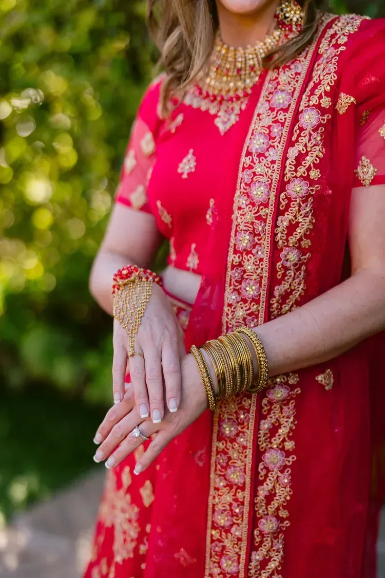 Woman in a bright pink embroidered traditional outfit with gold jewelry, standing outdoors with hands clasped.