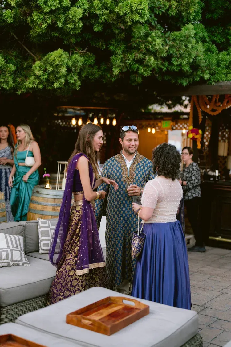 Guests chatting at an outdoor party under trees, with tables, seating, and warm string lights.