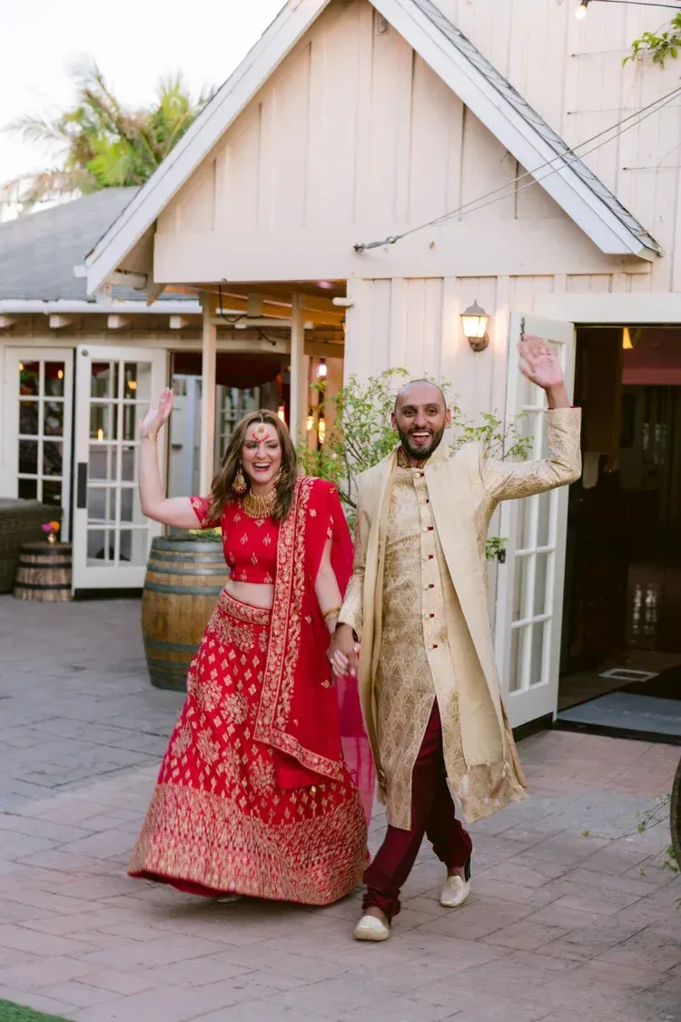 Couple in red and gold traditional attire walking outside a white house, smiling and holding hands