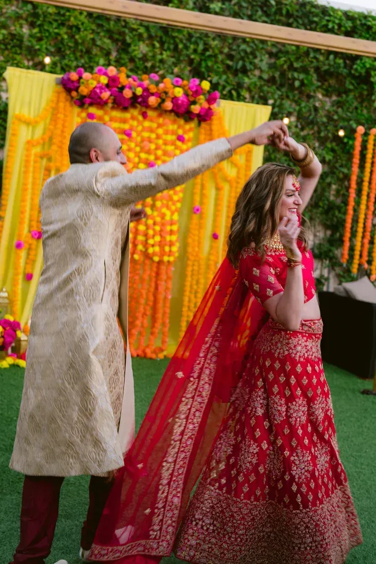 Couple dancing under colorful floral decorations, with the woman in a red lehenga and the man in a beige sherwani