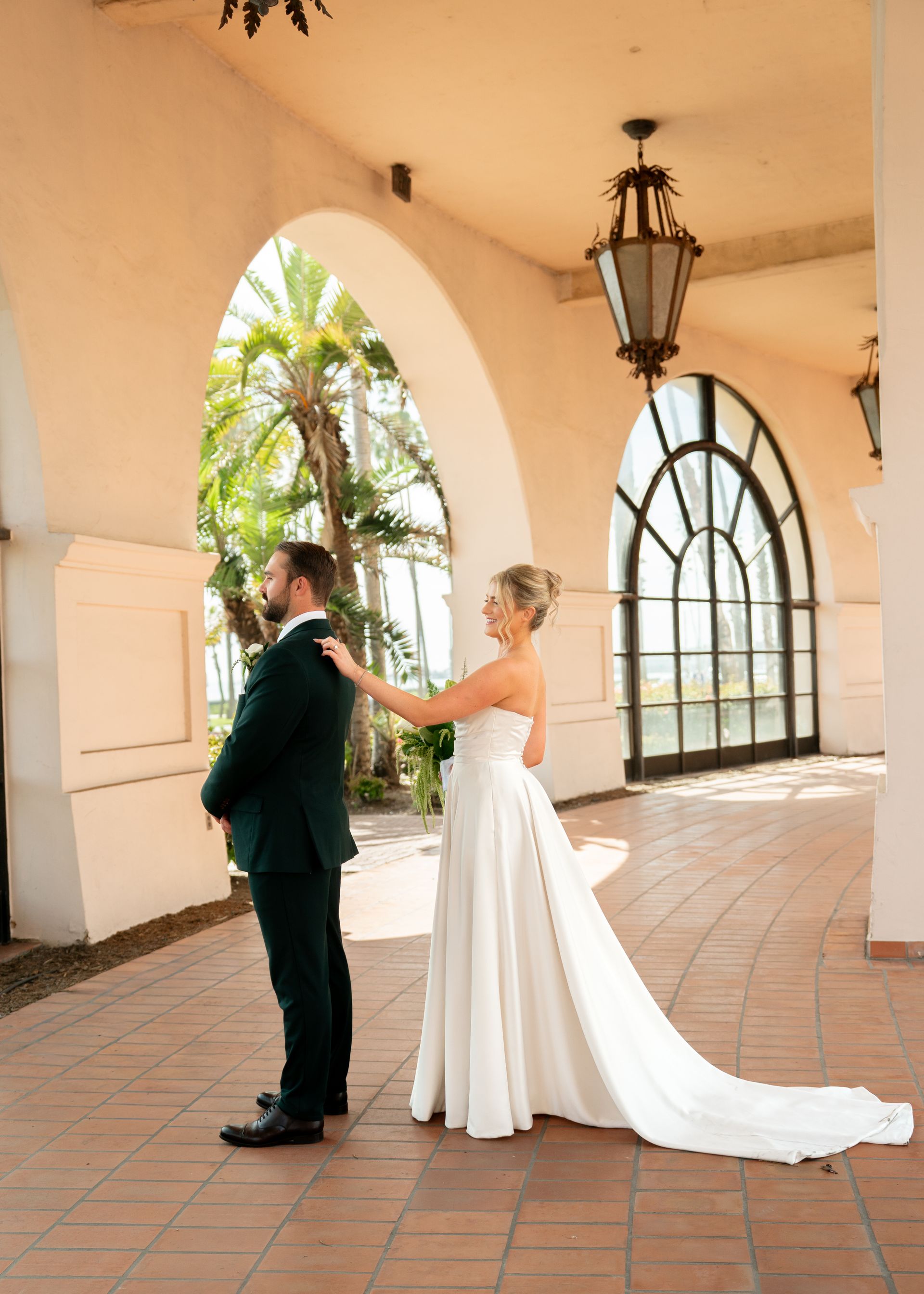 Bride and groom holding hands in a sunlit arched courtyard, with the bride’s long white gown trailing behind.