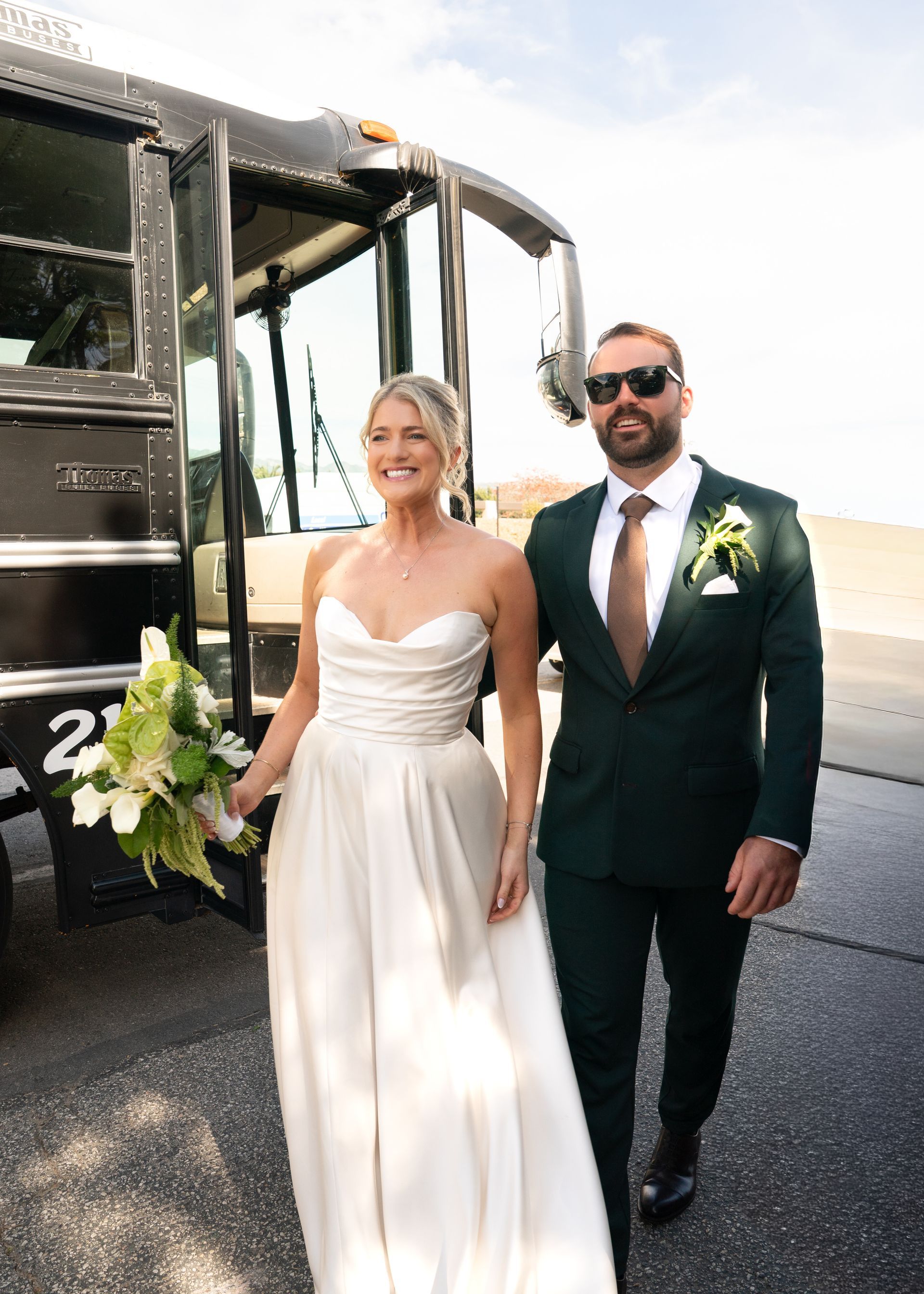 Bride in a white gown walks beside groom in a dark suit near a decorated bus outdoors