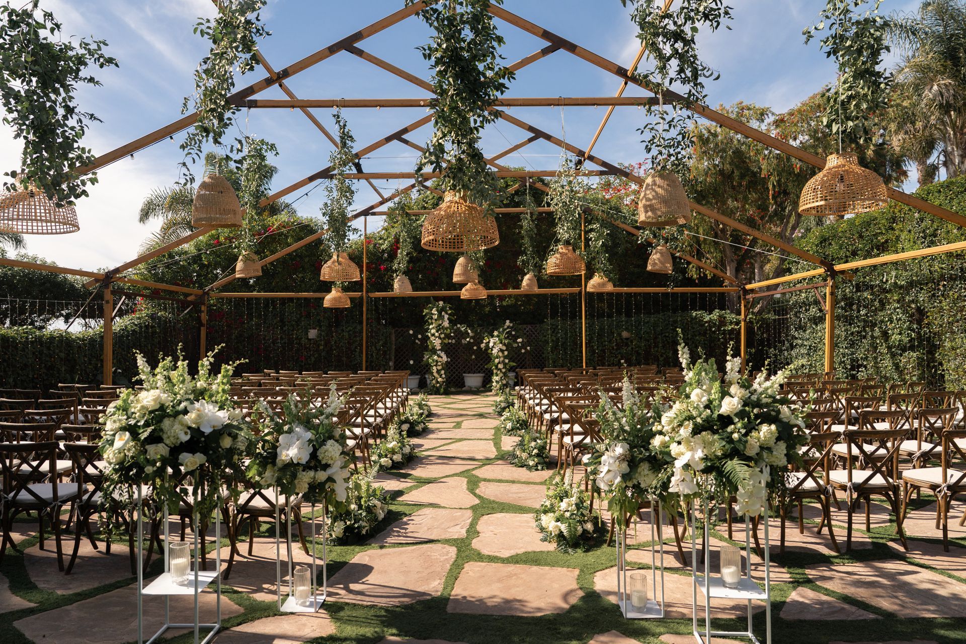Outdoor wedding ceremony setup with flower-lined aisle, chairs, and rustic greenery arch under a wooden canopy