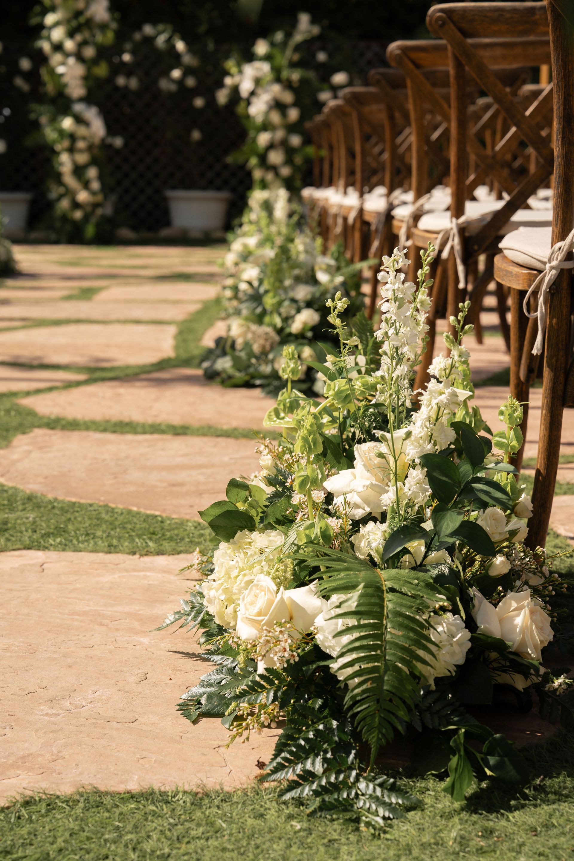 White floral arrangements line a garden aisle beside wooden chairs at an outdoor ceremony