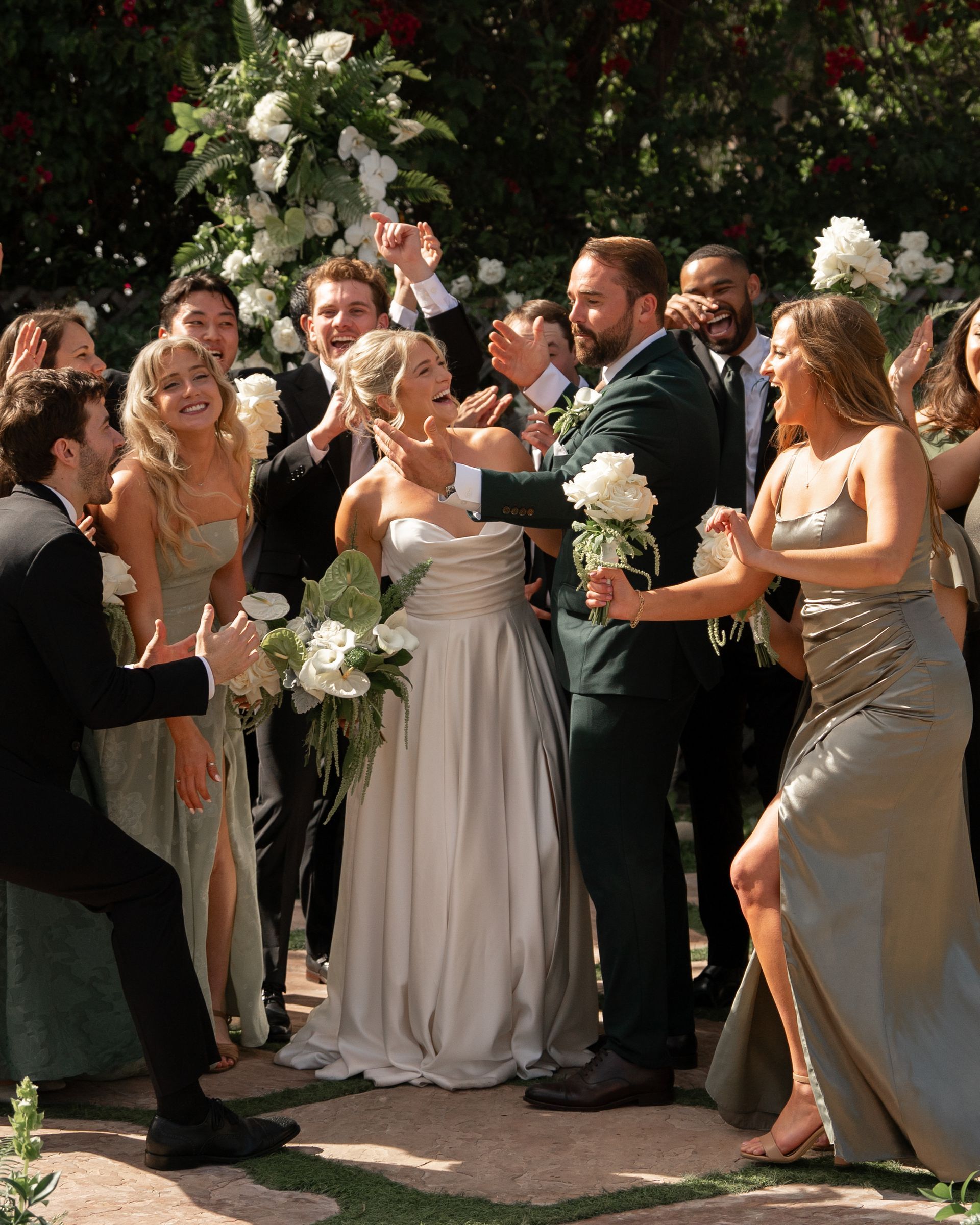 Guests celebrate at an outdoor wedding, clapping and holding white bouquets around a couple.