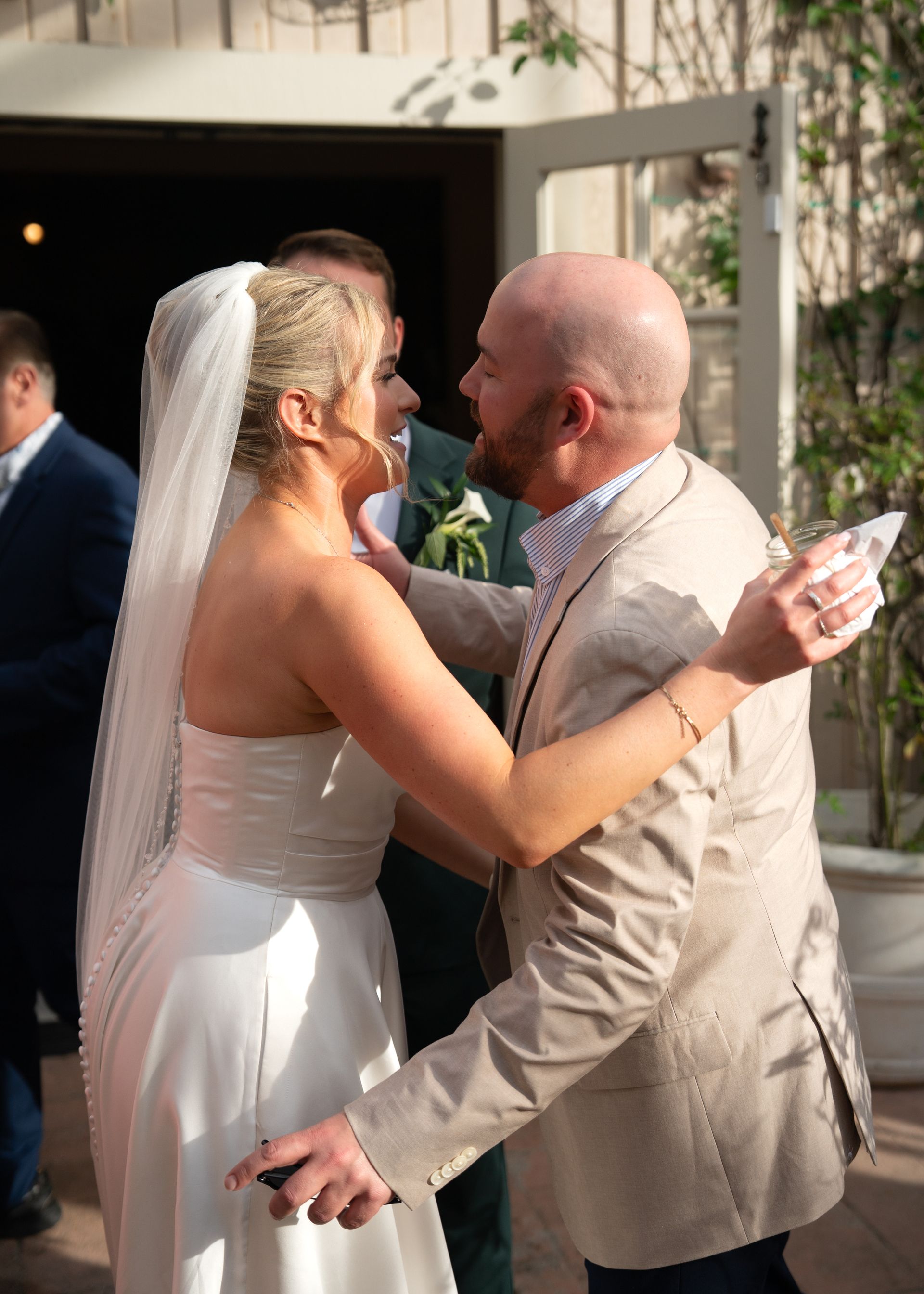 Bride in a white dress and veil embraces a man in a beige suit at a wedding