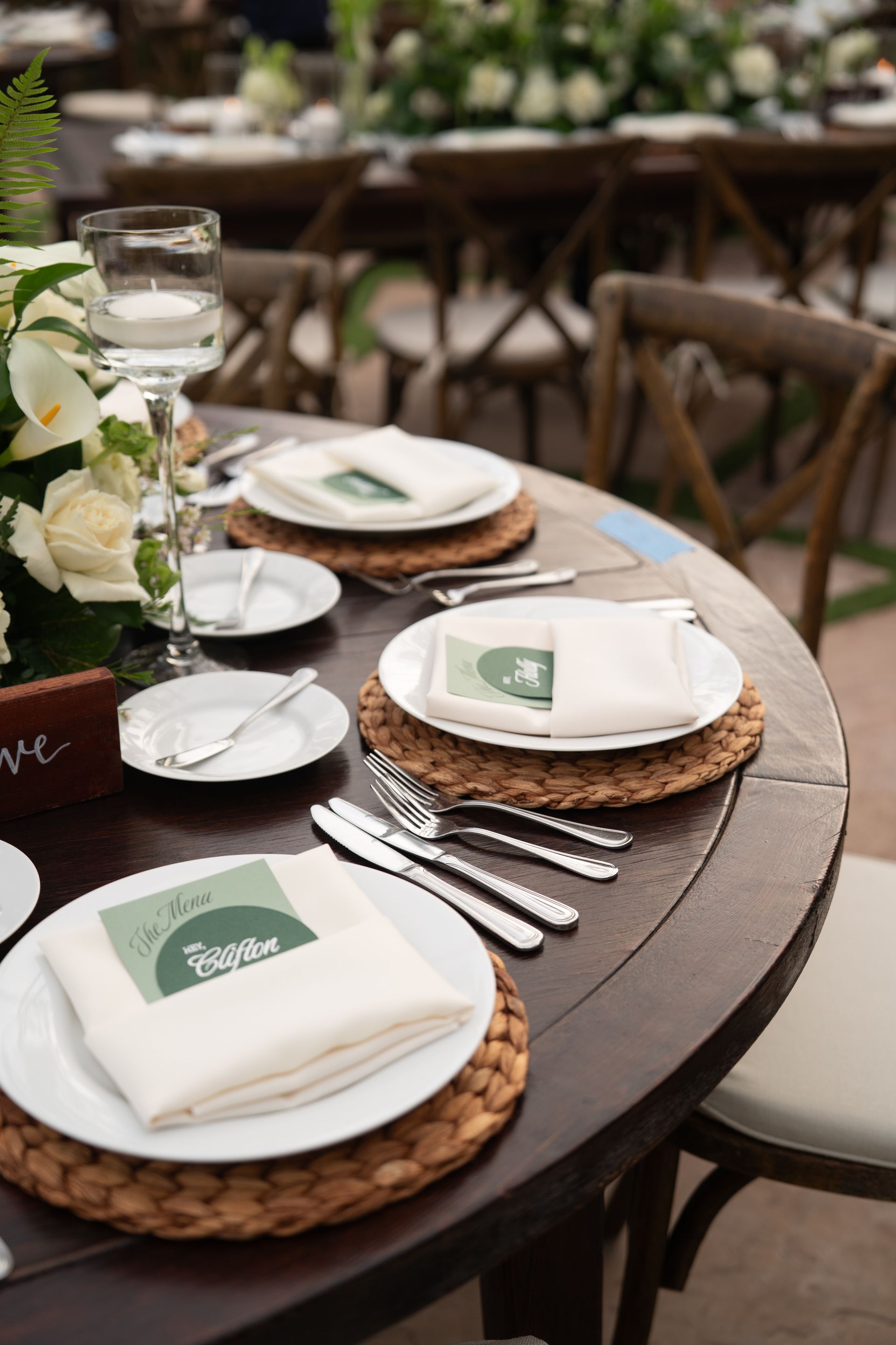 Elegant outdoor table setting with white plates, woven chargers, silverware, and glasses on a dark round table